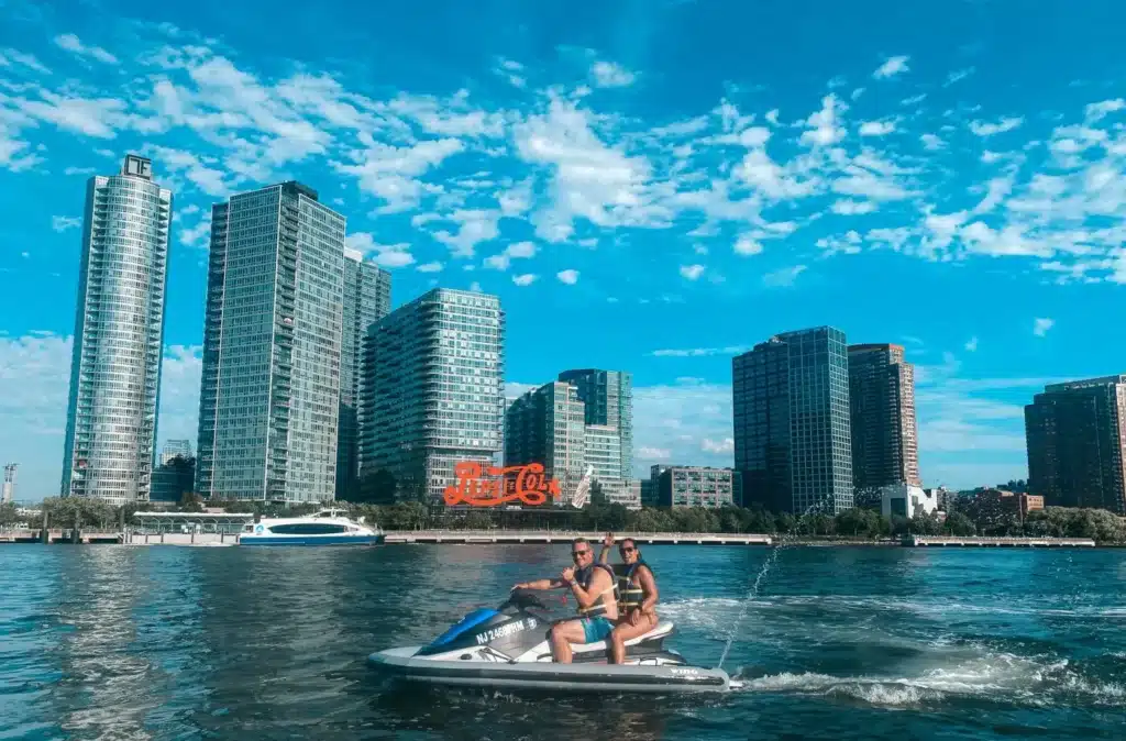Riders on a jet ski in New York Harbor with waterfront skyscrapers behind them
