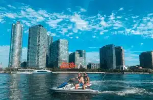 Riders on a jet ski in New York Harbor with waterfront skyscrapers behind them
