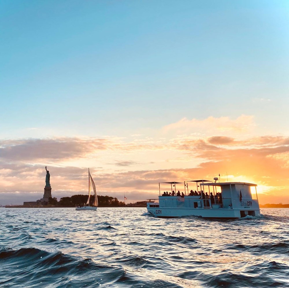 Boat near Statue of Liberty at sunset with sailboat, sky, and water.
