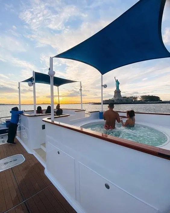 People in a hot tub on a boat with the Statue of Liberty in the background during sunset.